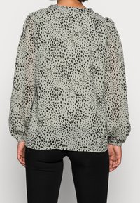 Genomskinlig grön blus med svart prickig mönster, puffärmar, rund halsringning och en slät yta, parat med svarta åtsittande byxor.