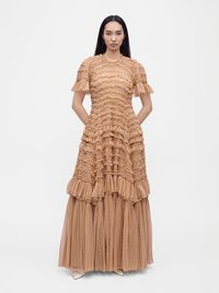 Femme debout, les mains dans le dos, portant une longue robe beige à volants avec des manches courtes et une jupe transparente superposée.