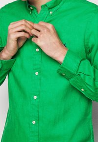 Grön skjorta med knappar i texturerad tyg med synlig sömnad och en liten logotyp. Har krage och knappmanschetter.