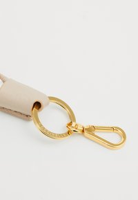 Porte-clés en cuir beige avec une texture lisse, doté d'un anneau et d'un fermoir en métal doré. Logo gravé sur l'anneau.