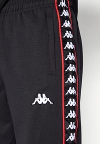 Μαύροι φόρμες με κόκκινες ρίγες στο πλάι, διακοσμημένοι με επαναλαμβανόμενο λευκό λογότυπο. Κατασκευασμένοι από μαλακό ύφασμα με ελαστική μέση και πλαϊνές τσέπες.