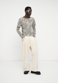 Cămașa gri cu camuflaj cu mâneci lungi se asociază cu pantaloni cremuți, lejeri. Modelul poartă pantofi negri masivi și ochelari de soare argintii.