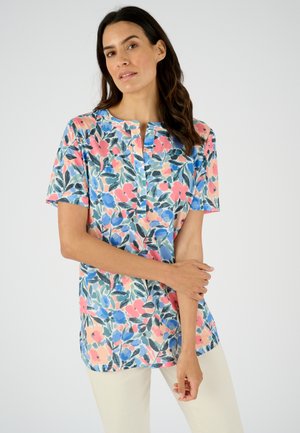 Donna che indossa una camicetta a maniche corte con motivi floreali nei toni del rosa, blu e verde, abbinata a pantaloni chiari, in piedi davanti a uno sfondo semplice.