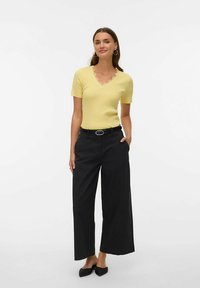 Geel geribd topje met korte mouwen en een gegolfde halslijn, gecombineerd met zwarte wijde broekspijpen en zwarte platte schoenen. Model staat met handen in de zakken.