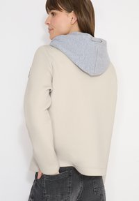 Femme portant une veste beige avec une capuche grise, mains dans les poches arrière d'un jean noir, tournée vers la gauche sur fond blanc.