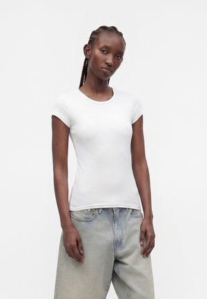 Ung kvinde med fletninger iført en tætsiddende hvid kortærmet skjorte og lysegrå løstsiddende jeans, stående mod en ensfarvet baggrund.