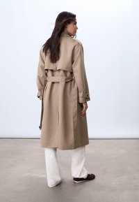 Trench coat beige con cintura, davanti doppiopetto e maniche lunghe. Presenta un'apertura sul retro e dettagli con bottoni. Indossato sopra pantaloni bianchi.