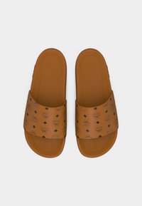 Sandalias marrones tipo slip-on con logos MCM negros y patrones de diamantes en las correas anchas, suela plana y plantilla texturizada, vistas desde arriba.