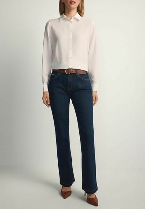 Kobieta ubrana w białą koszulę na guziki z długim rękawem, ciemnoniebieskie jeansy o prostych nogawkach z brązowym paskiem oraz brązowe szpiczaste buty na wysokim obcasie, stojąca nieruchomo.