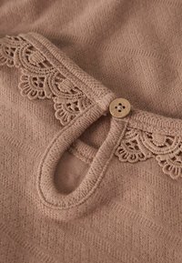 Vaaleanpunainen tekstiili, jossa on teksturoitu kuvio, koristeellinen pitsireunus ja puinen napin yksityiskohta lenkillisen aukon lähellä.