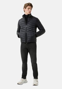 Schwarze Steppjacke mit horizontalem Steppmuster, Reißverschluss, hohem Kragen und zwei Seitentaschen. Kombiniert mit dunklen Jeans und weißen Sneakern.