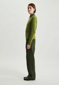 Pull en laine vert avec un ourlet côtelé, associé à un pantalon en velours côtelé vert foncé. Le mannequin porte des chaussures noires et une chemise à motifs sous le pull.