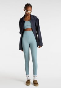 Lichtblauwe sportoutfit bestaande uit een cropped top en hoge taille legging, gecombineerd met een donkere shirt met knopen en tan sneakers.