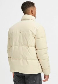 Chaqueta acolchada beige con cuello de canalé y detalles cosidos. La tela parece suave y lisa, con un acabado ligeramente brillante.