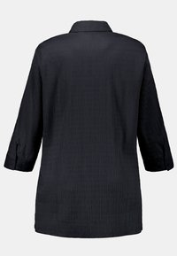 Maglietta nera con maniche a tre quarti, superficie testurizzata con linee verticali e colletto standard. I polsini abbottonati completano il design.