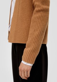 Brauner gestrickter Cardigan mit strukturiertem Rippdesign, verkürzter Länge und weitem Ärmel, kombiniert mit einem weißen Hemd darunter.