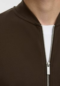 Brūns bumbvedēja jaka ar teksturētu virsmu, aprīkota ar augstu apkakli un sudraba rāvējslēdzēju. Zem tās redzama balta krekla.