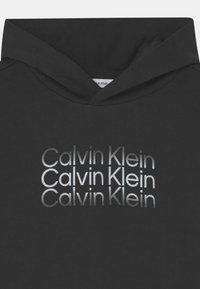 Sudadera negra con un diseño gráfico plateado de "Calvin Klein" en diferentes fuentes y tamaños, con un diseño estándar de capucha con cordón.