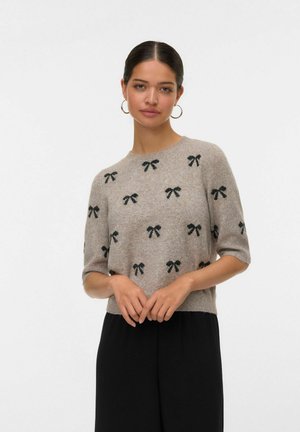 Hellgrauer Strickpullover mit kurzen Ärmeln, der mit schwarzen Schleifenmustern verziert ist und einen runden Ausschnitt hat. Kombiniert mit schwarzen Hosen.