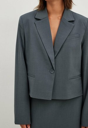 Femme portant un blazer gris foncé à un bouton avec revers crantés et jupe assortie sur un fond clair uni.