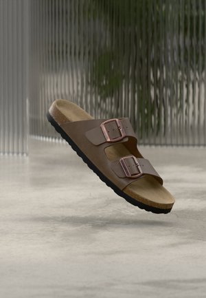 Sandalo marrone con due cinturini regolabili con fibbia e una soletta sagomata in sughero che sembra galleggiare sopra un pavimento in cemento.