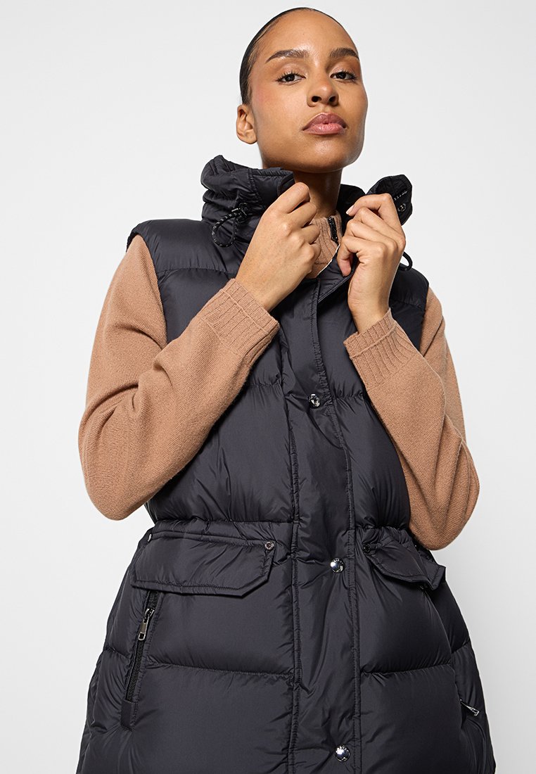 Schwarze Steppweste mit hohem Kragen, Reißverschluss vorne, Seitentaschen und taillierter Passform. Über einem beigefarbenen Strickpullover getragen. Glatte Textur.