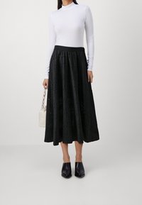 Larga falda negra con patrón de ojales, con cintura fruncida, llevada sobre un cuello de tortuga blanco, combinada con zuecos negros y un pequeño bolso blanco.