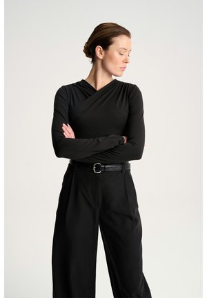 Kobieta z włosami związanymi w kok, ubrana w czarną bluzkę z długim rękawem i spodnie o szerokich nogawkach, stoi z założonymi ramionami, patrząc w lewo na jednolitym tle.