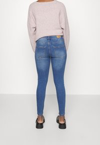 Person som bär en ljuslila tröja, blå högmidjade skinny jeans och svarta platåskor, stående mot en enkel vit bakgrund.