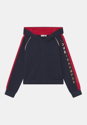 Marineblå lynlås-hættetrøje med røde striber på ærmerne, rødt hættens indre og gyldent tekst "LOVE IS A SUPER POWER" på højre ærme.