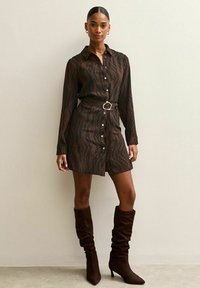 Mujer de pie contra una pared, vestida con un vestido camisero marrón con estampado de cebra y cinturón, y botas marrones de ante hasta la rodilla con un ajuste holgado.