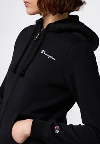 Sudadera con cremallera negra hecha de tela suave, con un logo blanco de "Champion" y un colorido detalle bordado en el puño.