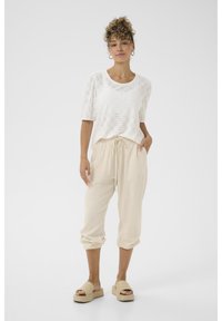 Blusa bianca in pizzo con maniche a sbuffo, abbinata a pantaloni beige larghi e sandali con zeppa beige. Tessuto morbido e vestibilità rilassata.