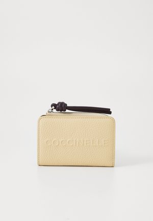 Portefeuille en cuir jaune avec une finition texturée, logo en relief, et un tirette de fermeture éclair violet foncé avec un détail en nœud. Forme rectangulaire compacte.