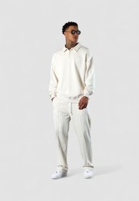 Helles Cremepullover mit einem Reißverschlusskragen und passenden Hosen, aus weichem Stoff gefertigt. Weiße Sneakers vervollständigen das Outfit.