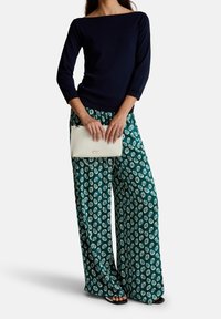 Marineblaues Langarmshirt mit Bootsausschnitt, kombiniert mit grünen weiten Hosen, die ein durchgängiges weißes Blättermuster aufweisen, sowie einer weißen Handtasche.