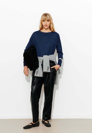 Haut à manches longues bleu marine avec texture mouchetée, pull gris noué à la taille, pantalons en simili cuir noirs, et chaussures noires à enfiler avec clous.