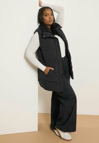 Svart pufferväst med knappframsidan, texturerad krage och sidofickor, bärs över en långärmad vit skjorta och vida svarta byxor.