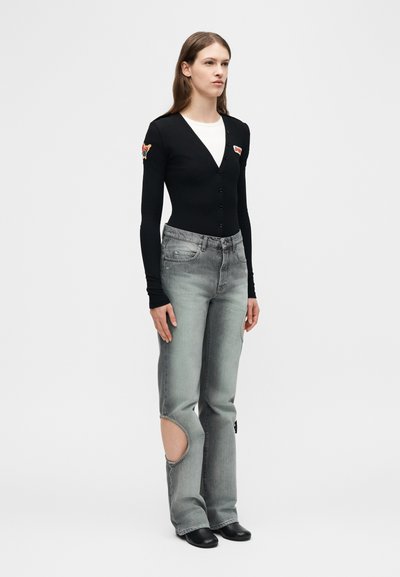 Femme debout portant un cardigan noir boutonné avec des patchs, une chemise blanche en dessous, un jean gris avec une découpe sur un genou, et des chaussures noires.