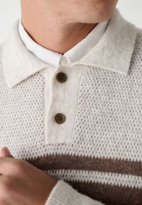 Svetlobéžový pletený sveter s textúrovaným vzorom, vybavený bielym golierom a tromi hnedými gombíkmi. Jemné hnedé vodorovné pruhy.