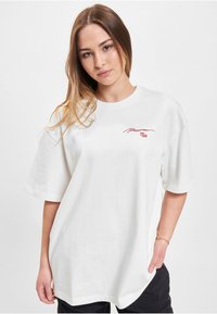 White oversized T-shirt made of cotton, featuring short sleeves, round neckline, and red embroidered logo on the chest.