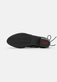 Baskets noires avec une semelle en caoutchouc texturé, présentant un design flexible et une marque rouge à l'arrière. Les lacets s'étendent de la zone de la languette.