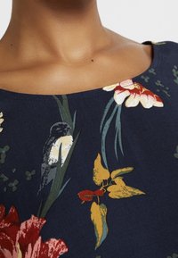 Collo e parte superiore del torace di una persona che indossa un top blu navy con un motivo floreale e di uccelli caratterizzato da colori rosso, giallo e verde.