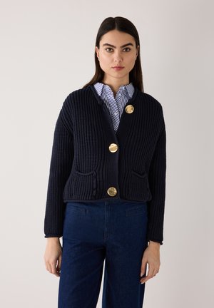 Woman wearing a navy knit cardigan with large gold buttons over a striped shirt and dark blue pants, standing against a plain background.