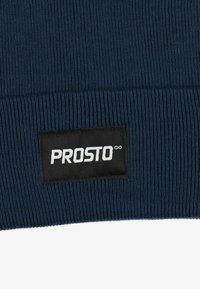 Granatowa prążkowana czapka typu beanie z czarną metką z tkaniny z białym logo "PROSTO" i symbolem nieskończoności, dodająca teksturę i detale marki.