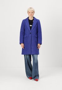 Purple textured coat with a notched lapel, single button closure, and side pockets, paired with wide-leg denim and red shoes.