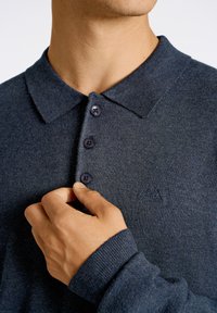 Mörkblå polo shirt med en strukturerad väv, tre lila knappar och en liten broderad logotyp på bröstet. Långärmad.