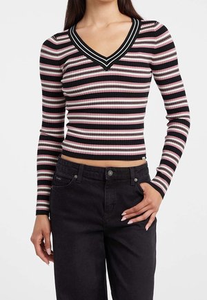 Kvinde iført en ribbet, langærmet cropped top med sorte, pink og hvide vandrette striber og V-hals, kombineret med sorte jeans.