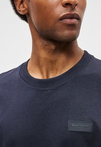 Κοντινή λήψη ενός άνδρα που φοράει ένα σκοτεινό μπλε μπλουζάκι με στρογγυλή λαιμόκοψη και μια μικρή μαύρη ετικέτα με το λογότ�υπο του Marc O'Polo στο στήθος.