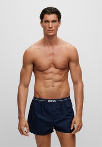 Navy blue boxer shorts with a button front and elastic waistband featuring the "BOSS" logo in white. Smooth texture and classic cut.